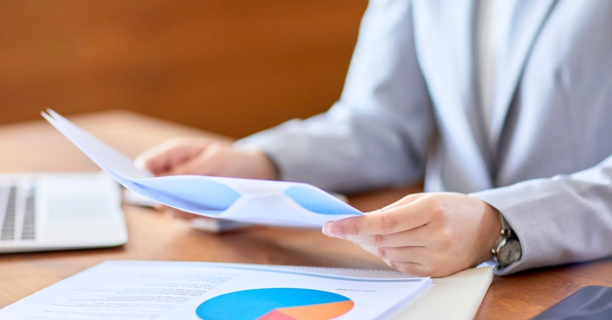 ビジネスシーンで、スーツを着た人物が書類を手に持ち、デスク上の資料やノートパソコンを見ながら作業している様子