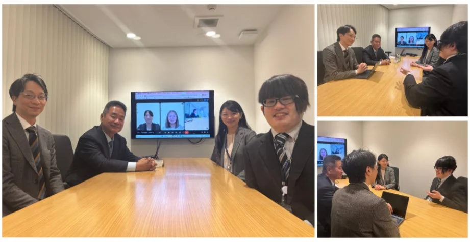 会議室で数人が対面で座り、大きなスクリーンにはオンラインで参加している人物が映し出されています。ビジネススーツを着た参加者たちが、活発な議論や情報交換を行っている様子が伺えます。ハイブリッド形式の会議の様子です。