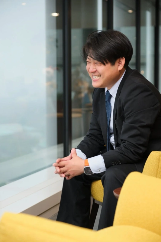 スーツを着たビジネスマンが窓際の黄色いソファに座り、満面の笑みを浮かべている