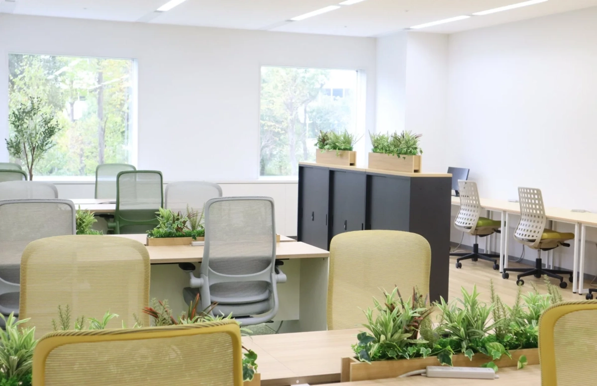 明るくモダンなオープンオフィスです。デスクには様々なデザインの椅子が並び、多くの観葉植物が配置されています。窓からは豊かな緑が見え、自然光が差し込む開放的で心地よいワークスペースです。