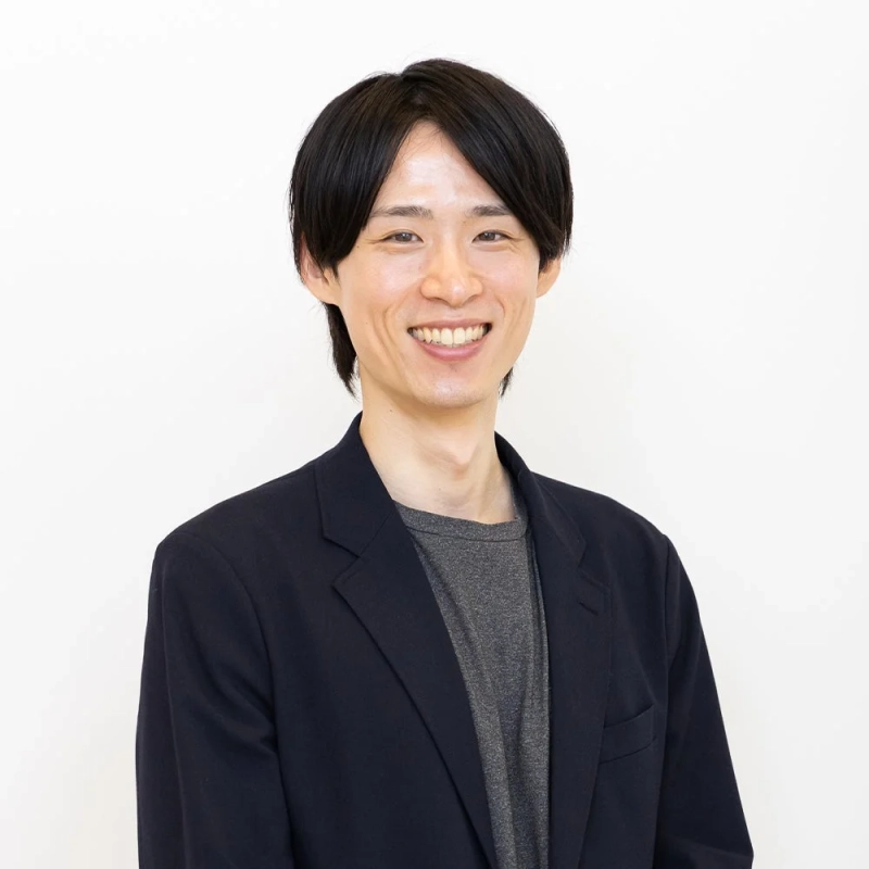 白い背景の前で、黒いジャケットとグレーのTシャツを着た若い日本人男性が、親しみやすい笑顔を見せている上半身のポートレート写真です。