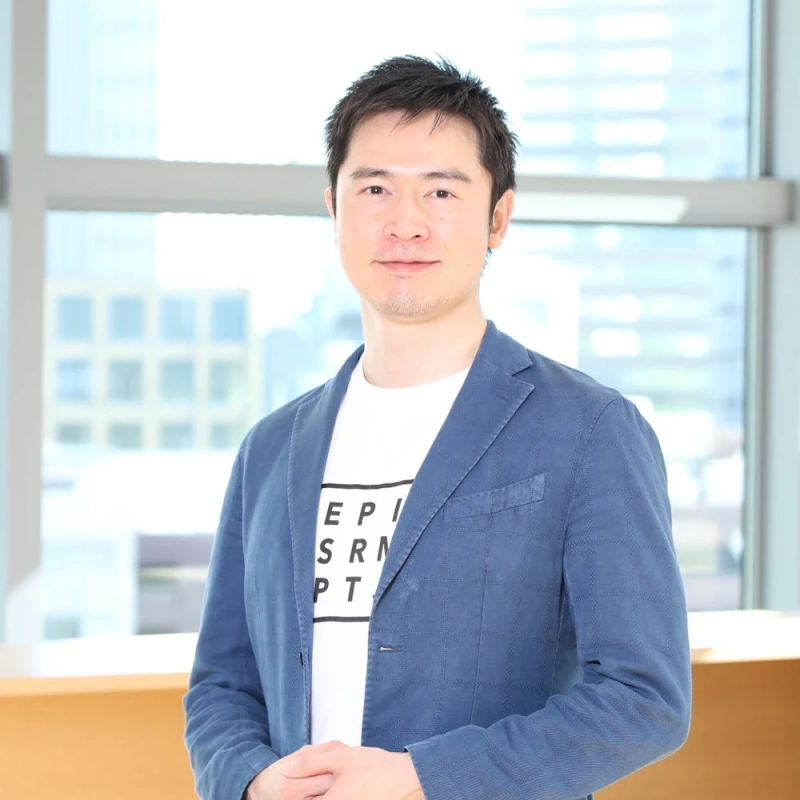 明るいオフィスを背景に、青いジャケットと白いTシャツを着たアジア人男性がカメラに向かって穏やかに微笑んでいます。