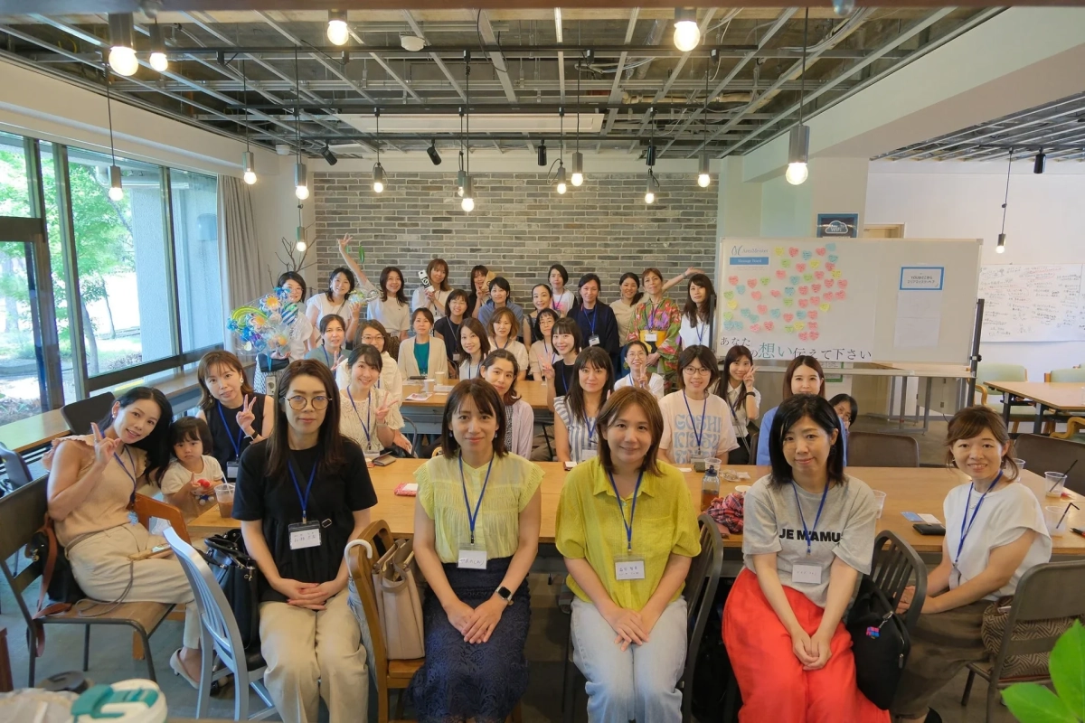 明るくモダンな空間で、多くの女性たちが笑顔で集合写真に収まっています。ワークショップかイベントの様子で、メッセージボードが背景に見えます。