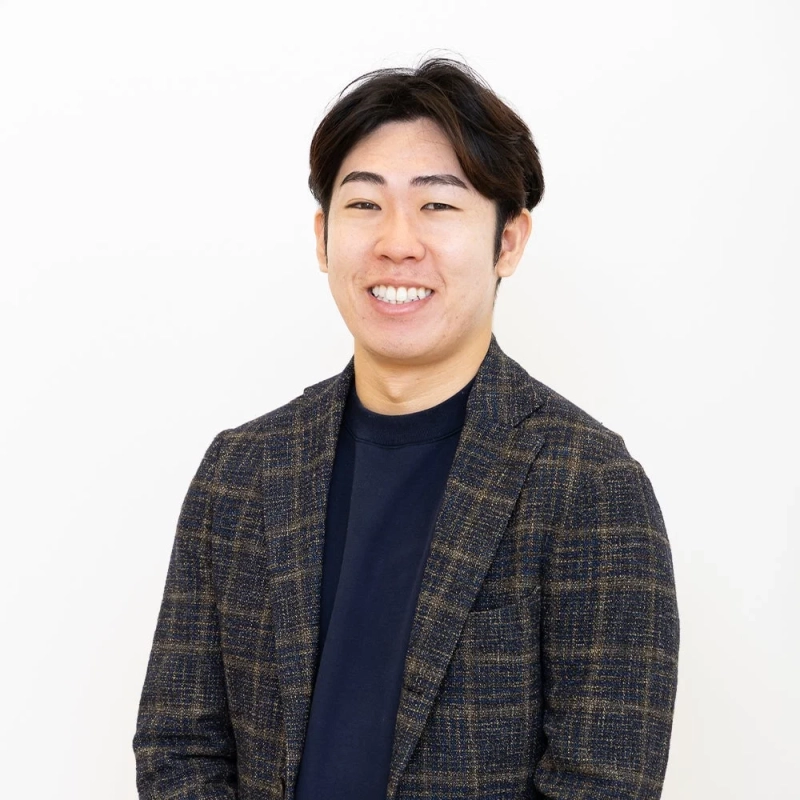 白い背景の前で、チェック柄のジャケットとネイビーのセーターを着た若い日本人男性が、カメラに向かって明るく笑顔を見せているポートレート写真です。