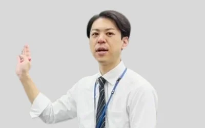 白いシャツとネクタイを着用した男性が、左手を上げて二本の指を立て、何かを説明しているような様子