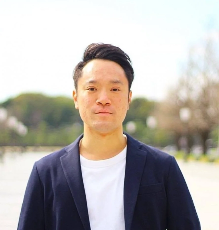 紺色のジャケットと白いTシャツを着用したアジア人男性のポートレートです。彼はカメラをまっすぐ見ており、背景にはぼかされた屋外の木々が見えます。真面目な表情で上半身が写し出されています。