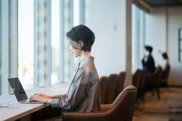 窓際に座り、ノートパソコンを使って仕事をする女性の横顔。オフィスやコワーキングスペースのようなモダンな空間で、集中して作業に取り組んでいる様子が伺えます。