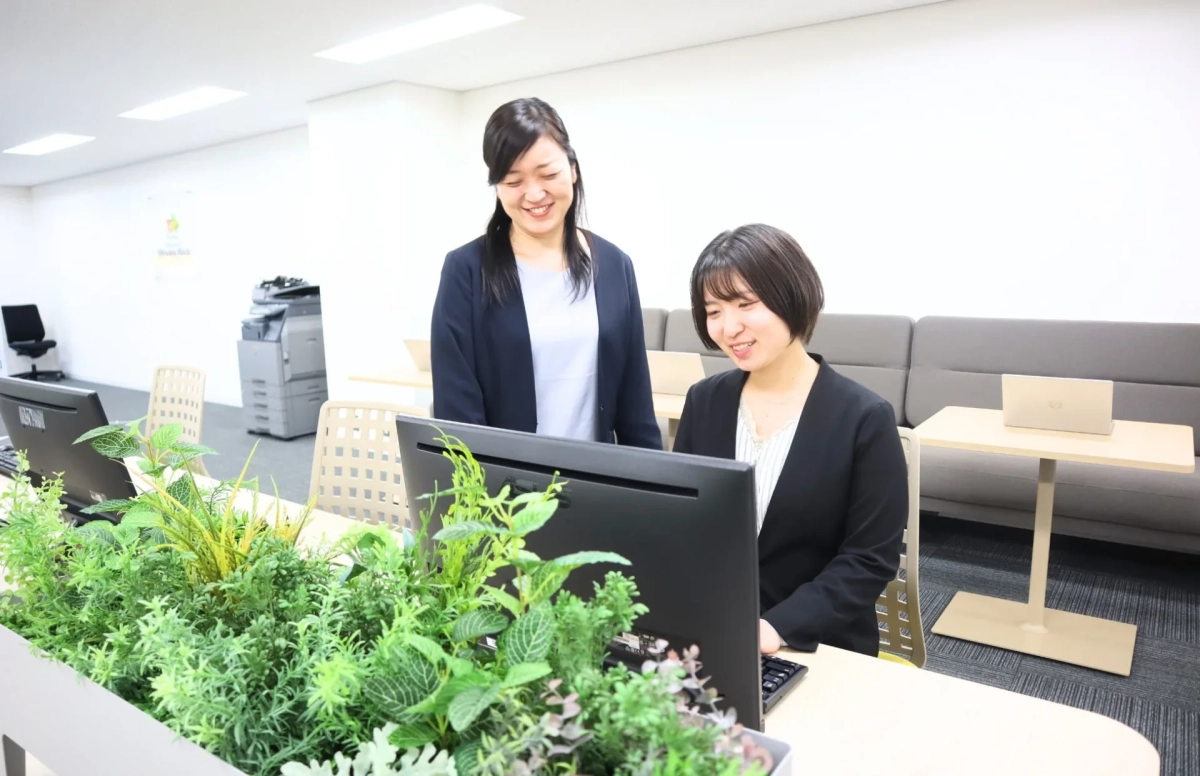 現代的なオフィスで働く2人の女性が描かれています。一人がパソコンに向かっており、もう一人が隣に立って画面を覗き込んでいます。二人とも笑顔で、和やかな雰囲気で業務に取り組んでいる様子がうかがえます。手前には観葉植物が配置されています。