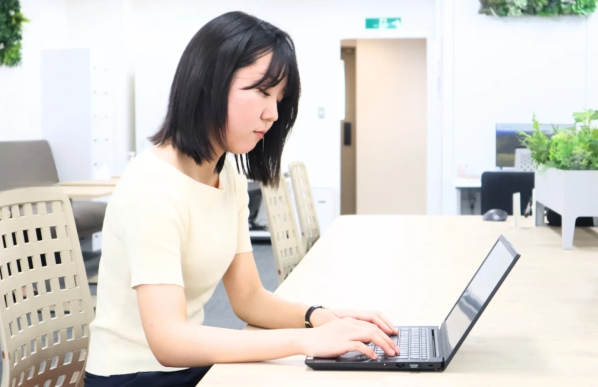 明るいオフィスで、若い女性がノートパソコンに向かい、真剣な表情で集中して作業をしている様子が写っています。