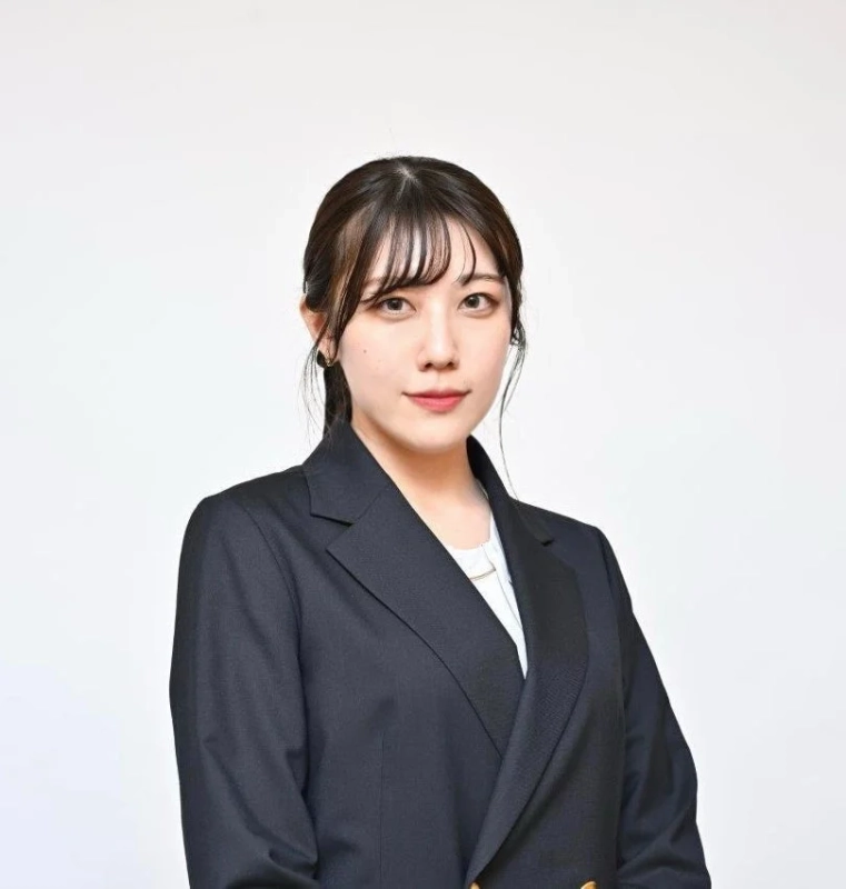 白い背景の前で、暗いスーツを着用した若い日本人女性がカメラをまっすぐ見ているポートレート写真です。