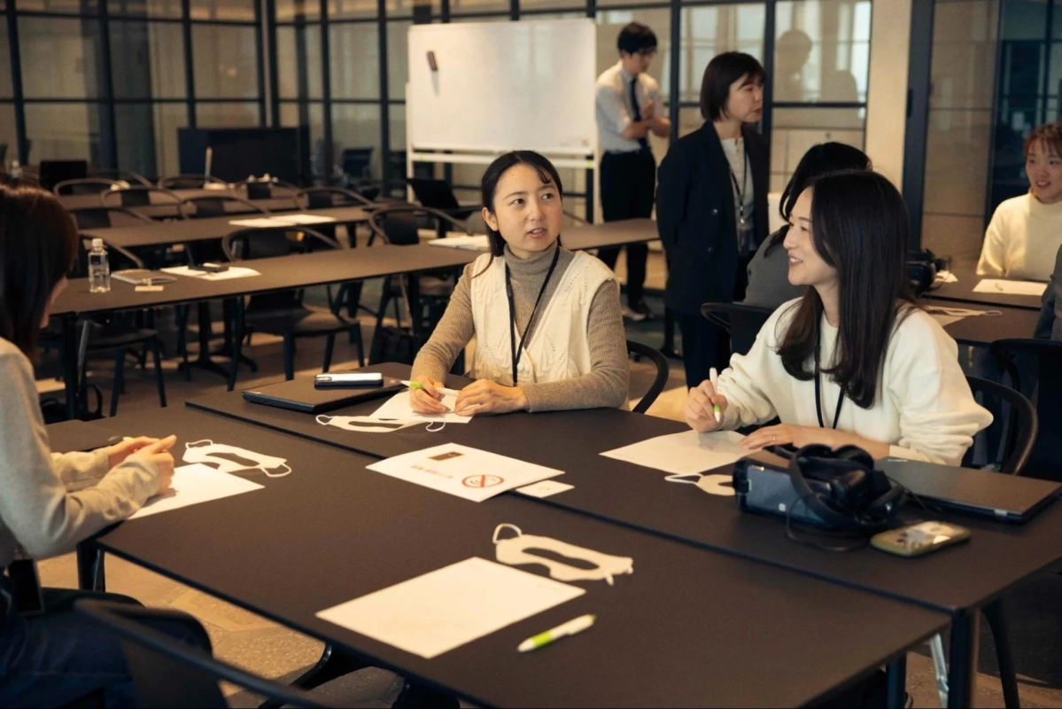 オフィスのような空間で、複数の人々がテーブルを囲んで座り、会議やセミナーに参加している様子。女性たちが中心となって活発に意見交換をしているように見え、集中した雰囲気の中で学習やディスカッションが行われている。