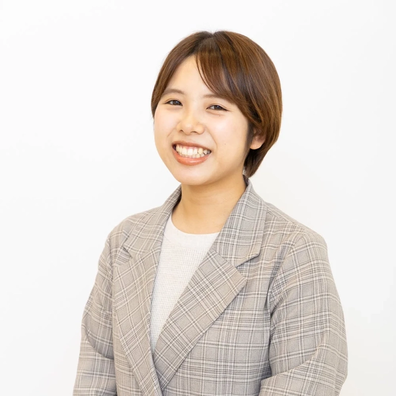 白い背景の前で、茶色のショートヘアの若い女性が明るい笑顔を見せています。グレーのチェック柄ジャケットと白いセーターを着用しており、親しみやすく清潔感のある印象です。