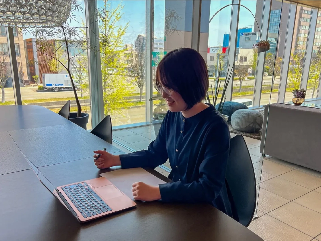 窓から街並みが見えるモダンな空間で女性がノートパソコンとペンを使って作業している様子