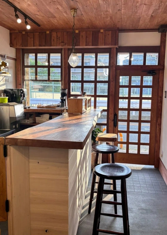 木の温もりが感じられるカフェの店内風景。長い木製カウンターとスツールが並び、格子窓から差し込む柔らかな自然光が心地よい空間を演出しています。