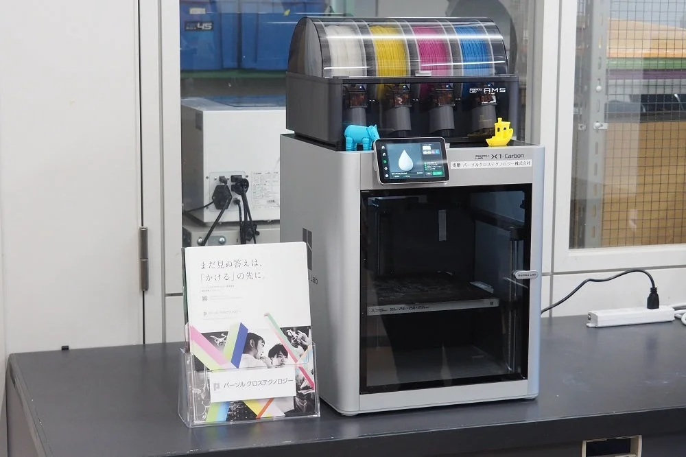Bambu Lab X1-Carbon 3Dプリンターがカラフルなフィラメントと共に設置されており、その隣にはパーソルクロステクノロジーのパンフレットが置かれています。研究開発またはオフィス環境での使用風景を示しています。