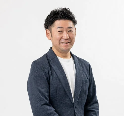 白い背景の前で、ダークブルーのジャケットと白いTシャツを着用したアジア人男性が、笑顔でカメラを見ている上半身のポートレート写真です。