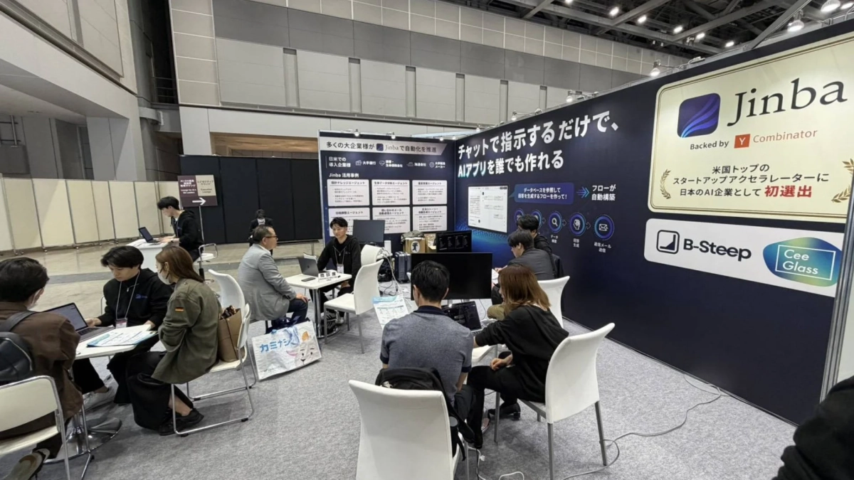 展示会場の「Jinba」ブースで、来場者がスタッフとAIアプリ開発プラットフォームについて商談しています。Y Combinatorに選出された実績や、チャットでAIアプリを簡単に作成できる機能が紹介されています。