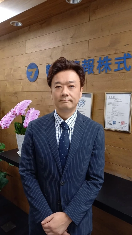 木目調の壁と企業ロゴ、胡蝶蘭があるオフィスのような場所で、ストライプのスーツを着た日本人男性が正面を向いて立っている