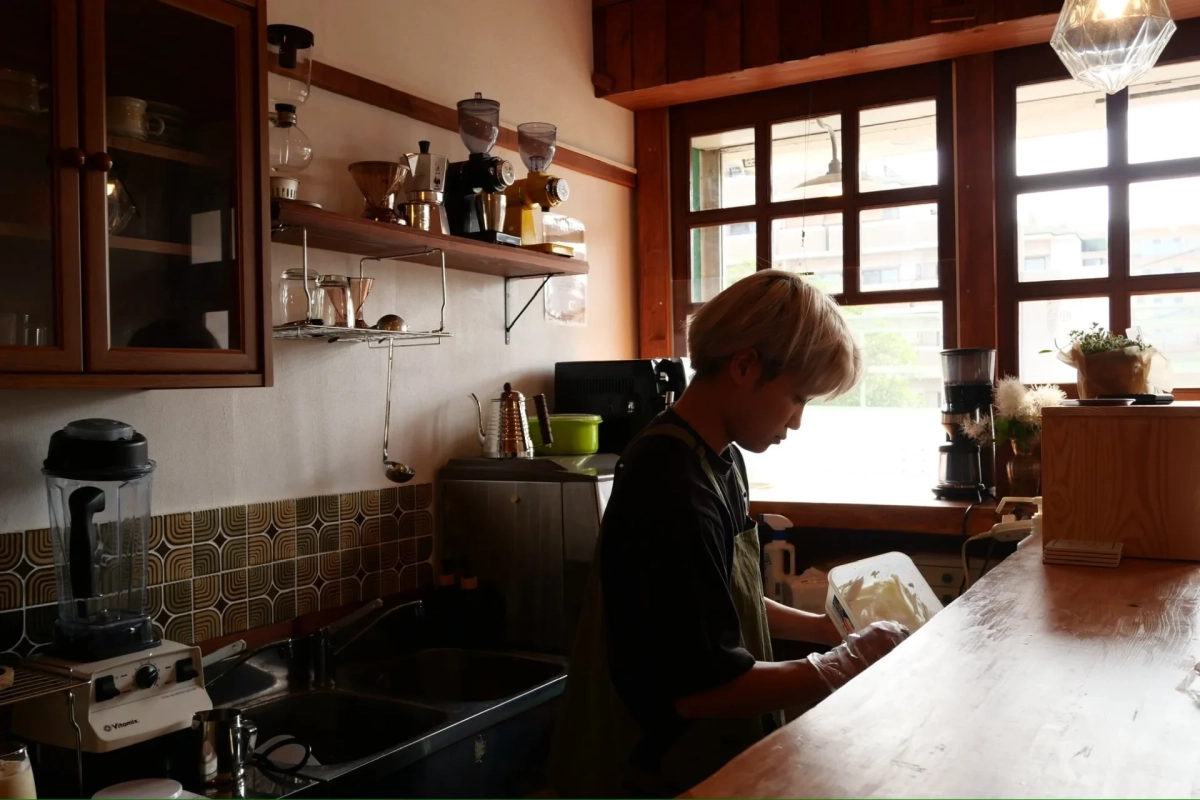 カフェの厨房で、エプロンを着用した男性が飲み物の準備をしている様子。コーヒーミルやブレンダーなどの器具が並び、温かみのあるレトロな雰囲気の空間です。