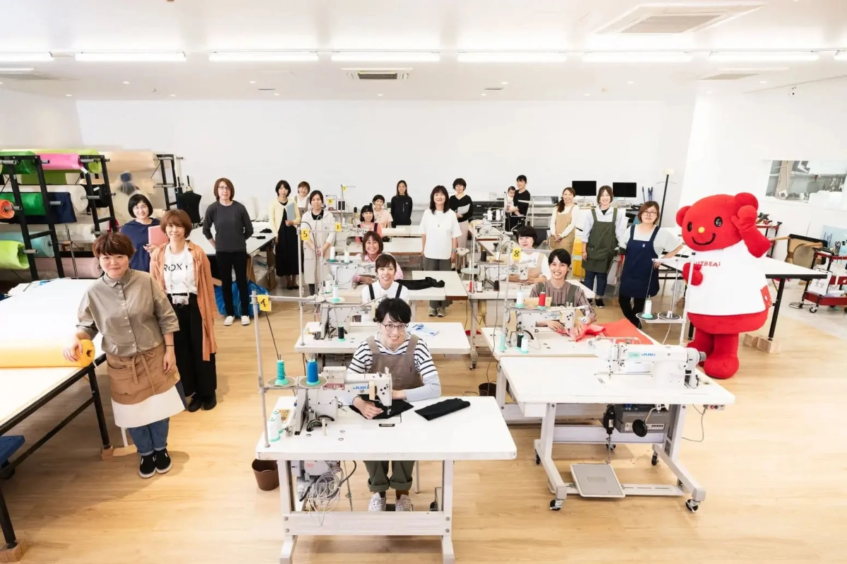 明るく広々とした縫製作業場で、多くの女性従業員がミシンと共に笑顔でポーズをとっている
