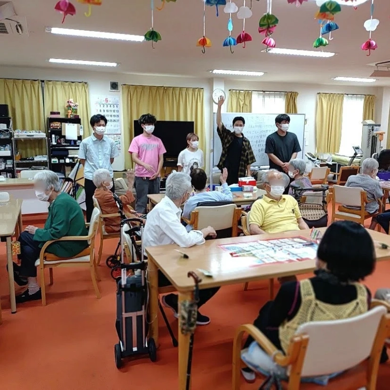 カラフルな傘の装飾が施された室内で、数名の若者が高齢者たちと交流している様子