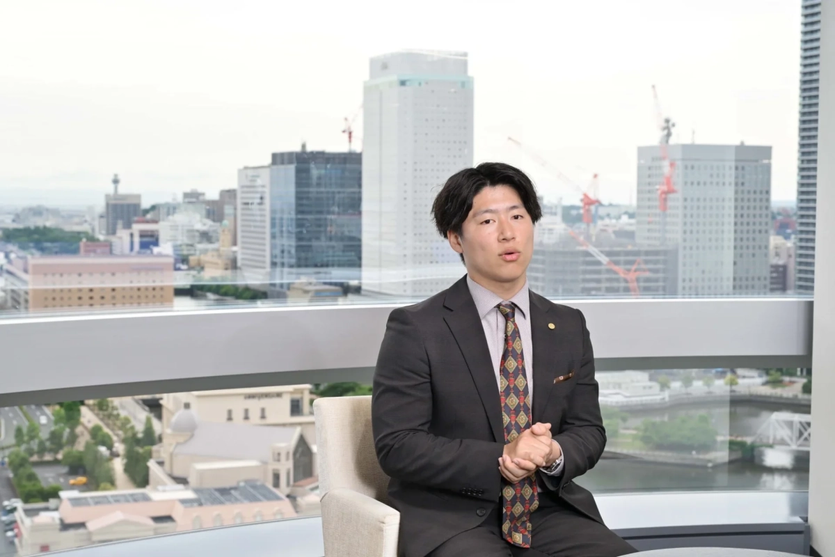 スーツ姿の男性が、高層ビル群と水辺の街並みを望む窓際で座り、話している様子。