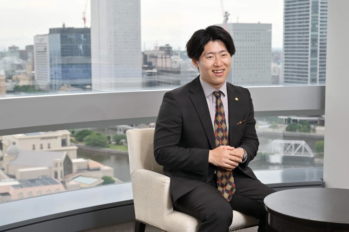 スーツを着た男性が、都市の景色を望む大きな窓際に座って笑顔を見せています。