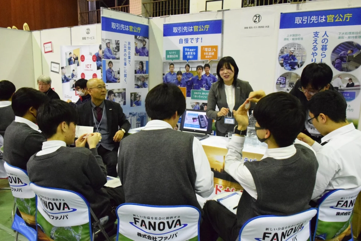 就職説明会のようなイベントで、複数の学生が企業ブースの担当者から説明を受けている様子