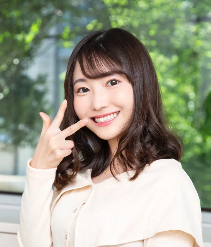 満面の笑みを浮かべた若い女性が、ピースサインのように指で口元を指しているポートレート。背景には緑の葉がぼやけて写っており、明るく爽やかな印象を与えます。