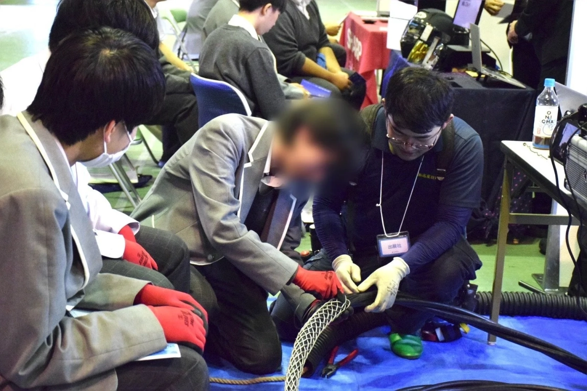 複数の人々が参加する技術実演または研修の様子。手袋をはめた二人が太いケーブルの作業を行っている
