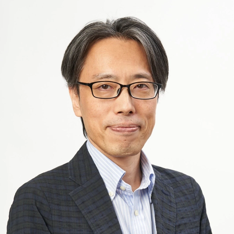 白い背景を背にした、眼鏡をかけたアジア系男性のポートレート。グレーの混じった髪とストライプのシャツ、柄物のジャケットを着用し、穏やかな表情をしています。