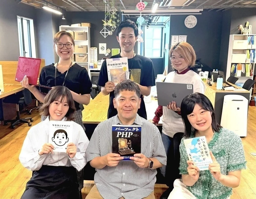 オフィスで働く6人の男女が笑顔でカメラにポーズをとっています。それぞれデザイン、プログラミング、動画編集に関する書籍やノートPCを持っており、チームの学習意欲や専門性を感じさせる一枚です。