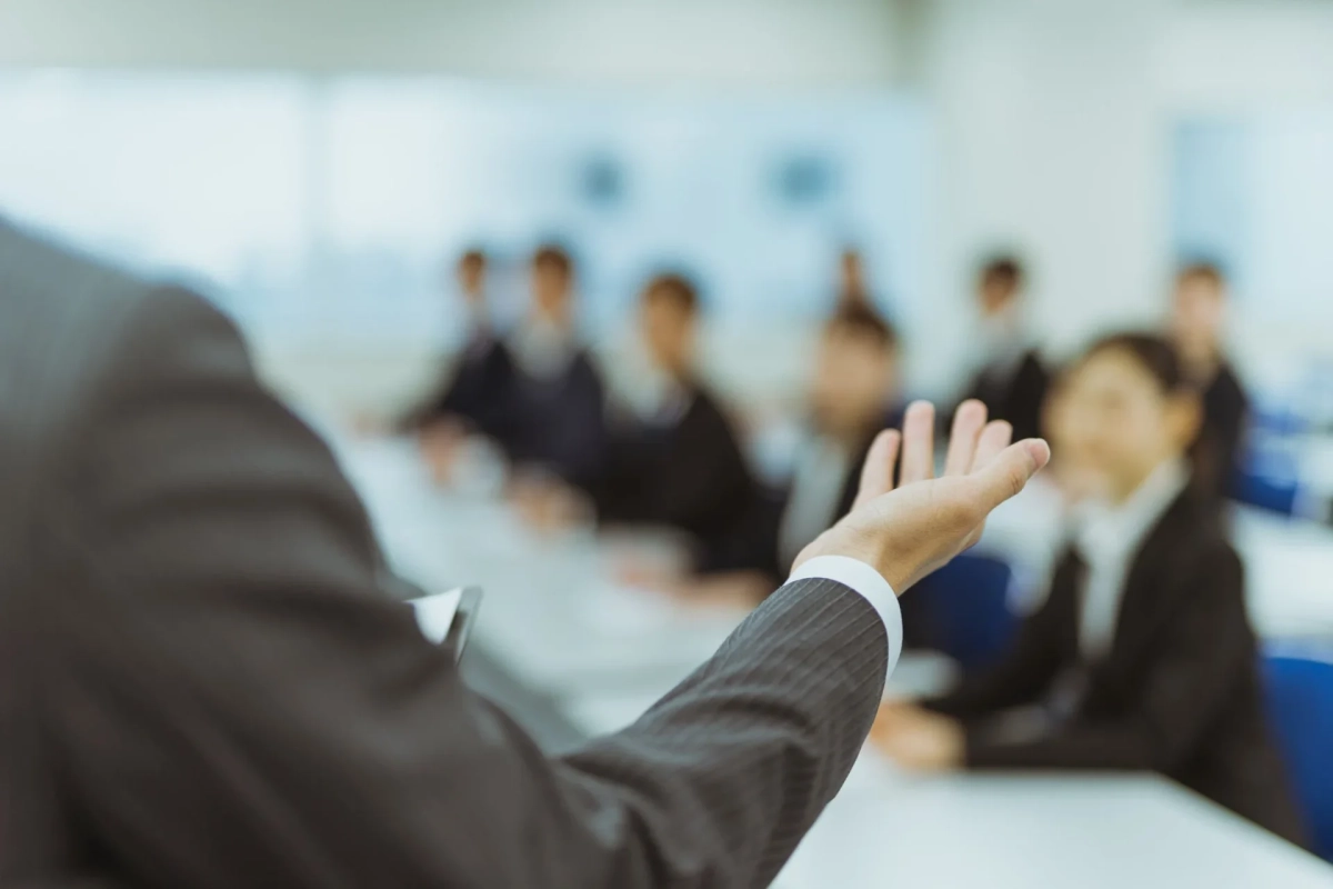 ビジネスシーンで、スーツを着た人物が聴衆に向かって手を広げ、プレゼンテーションや質疑応答をしている様子です。会議室やセミナー会場のような場所で、複数の参加者が座っています。