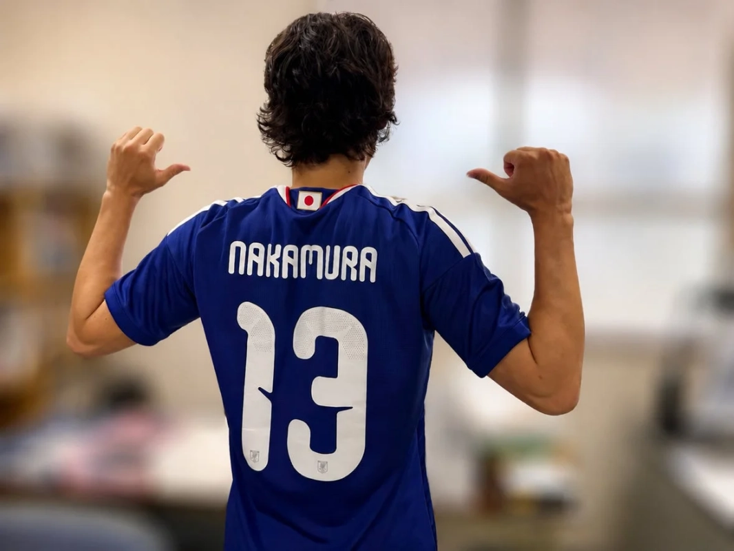 背番号13番、名前「NAKAMURA」と書かれた青いサッカー日本代表ユニフォームを着た人物の後ろ姿。両手の親指を上げてポーズをとっている。