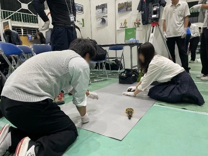 学生らしき2人が白い手袋を着用し、床に広げられたマットの上で小型のロボットを操作している様子