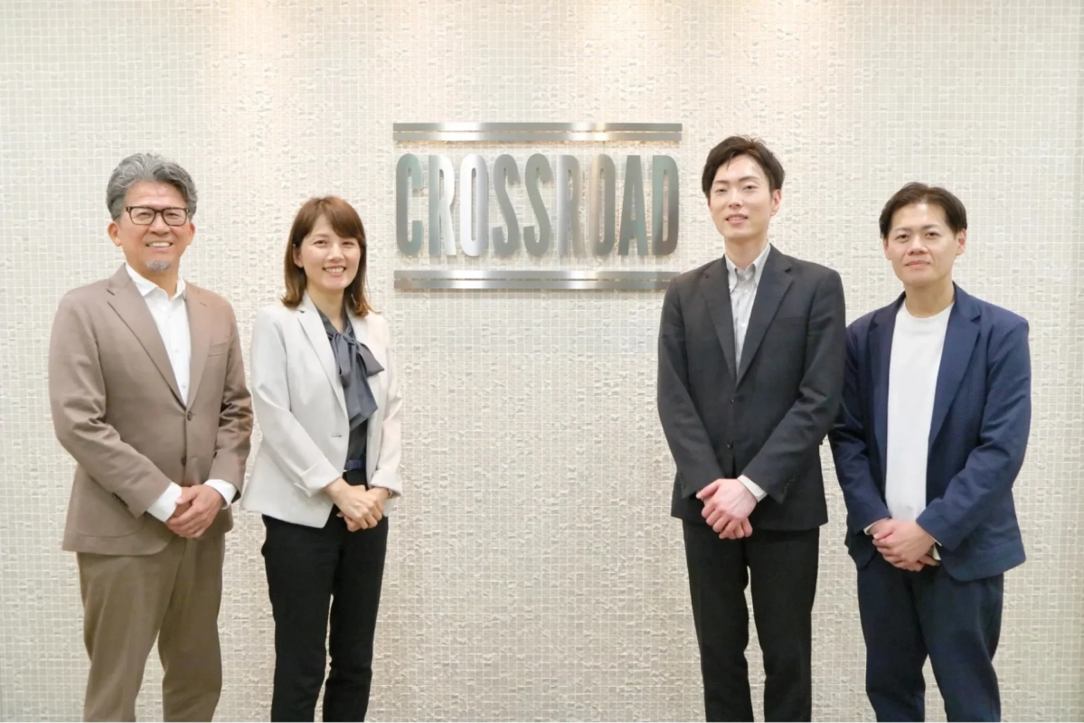 「CROSSROAD」のロゴがある壁の前で、スーツやビジネスカジュアルな服装の男女4人が笑顔で並んで立っている集合写真