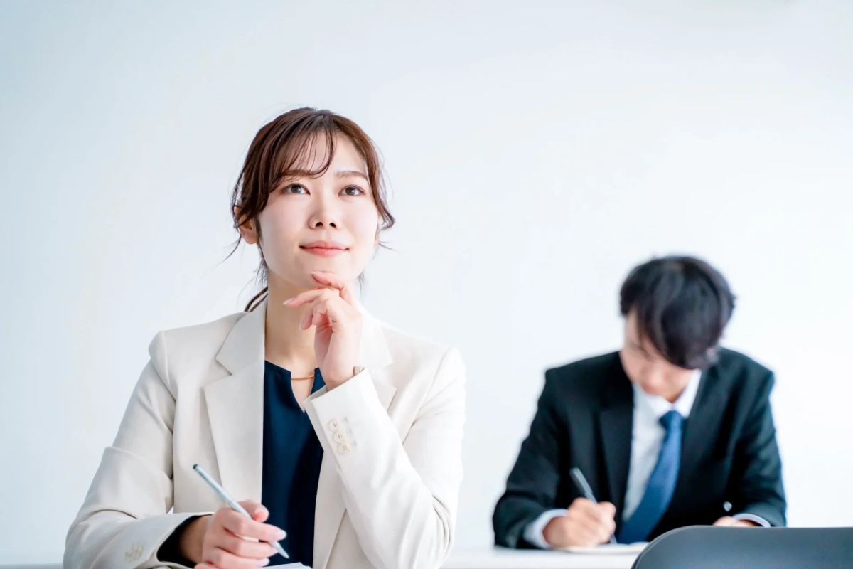 ビジネスシーンで、女性がペンを持ちながら何かを考え込むように上を見上げています。後方には別の男性がメモを取っており、会議やセミナーの様子を捉えた一枚です。