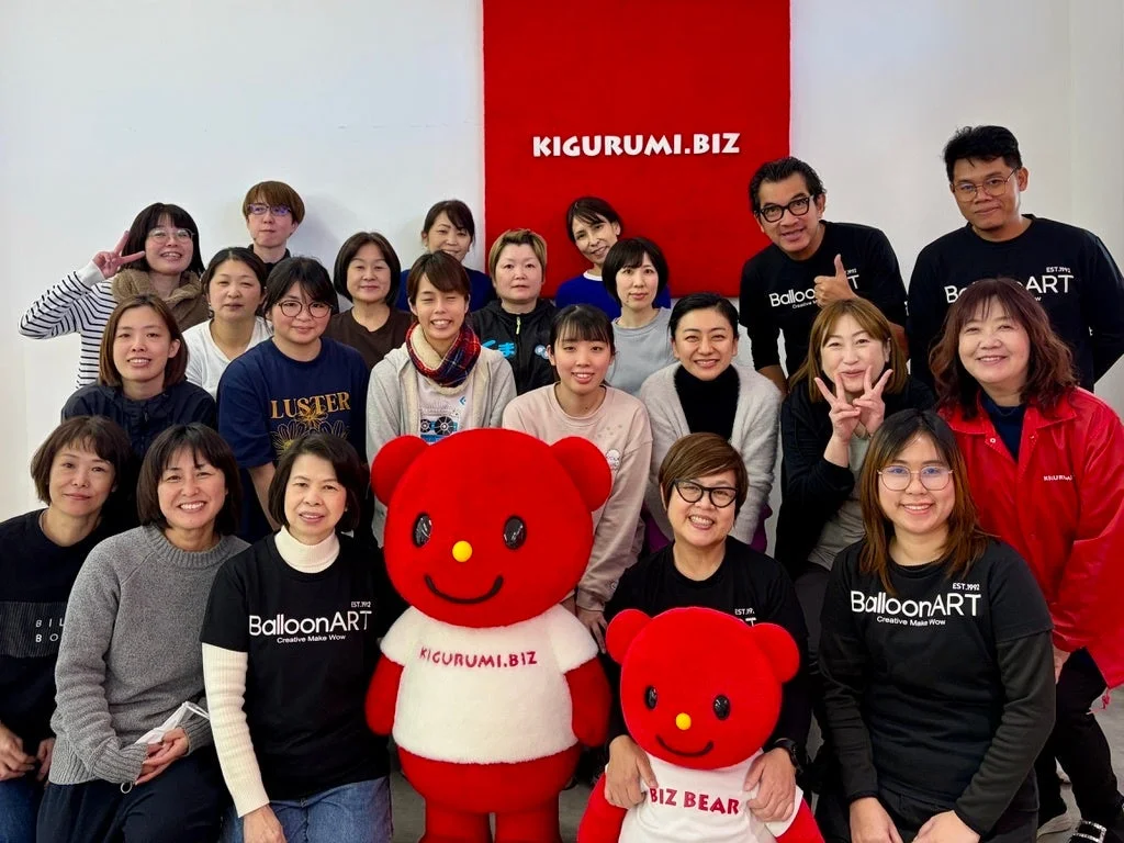KIGURUMI.BIZとBalloonARTのロゴがある赤い壁の前で、大勢の笑顔の男女が集合写真を撮っている