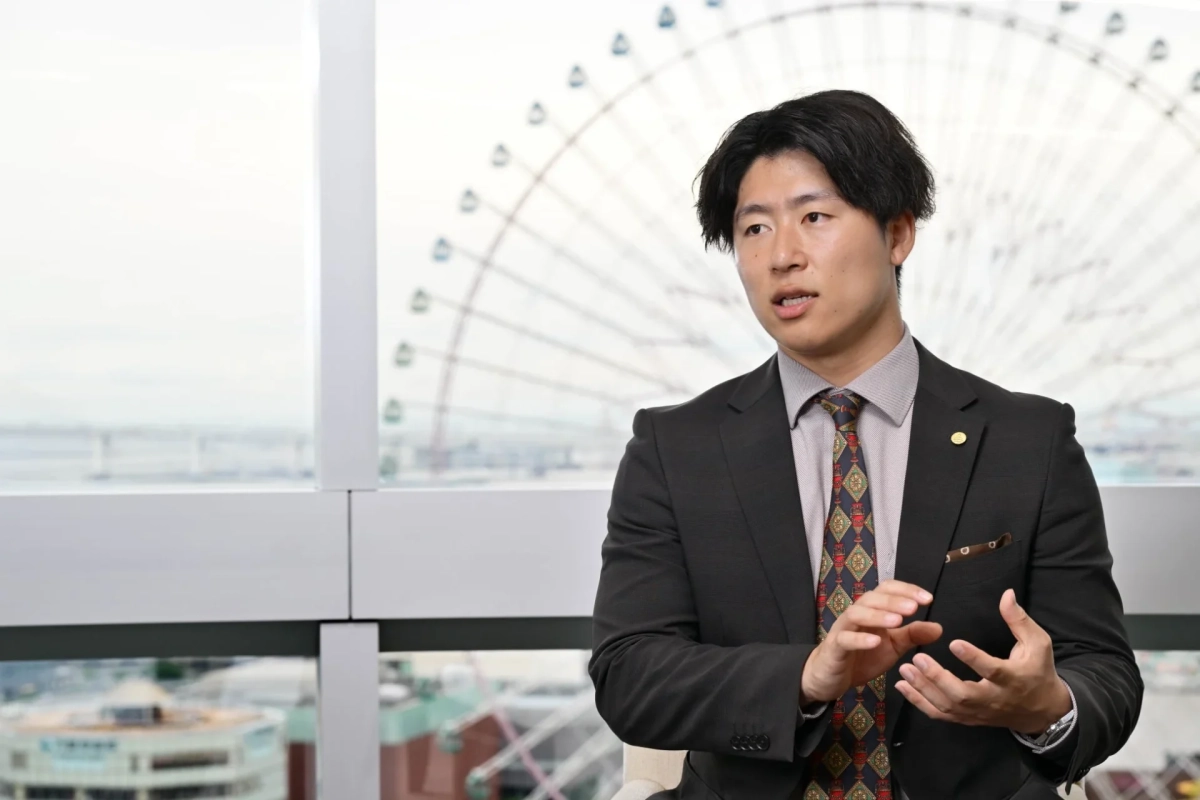 スーツを着た男性が窓際に座り、手でジェスチャーをしながら話している様子。