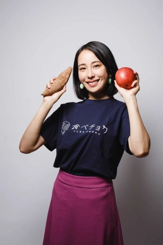笑顔の女性がサツマイモとリンゴを両手に持ち、「食べチョク」のロゴ入りTシャツを着て立っているスタジオ写真です。
