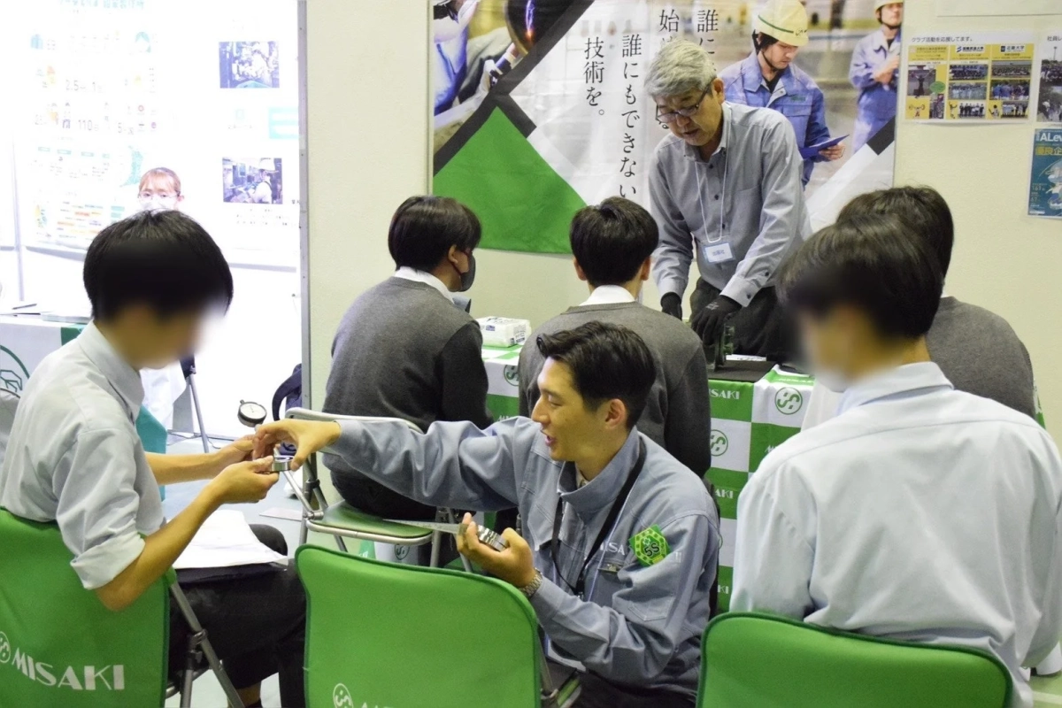 学生や大人がブースに集まっている様子。作業服の男性が何かを説明・実演している