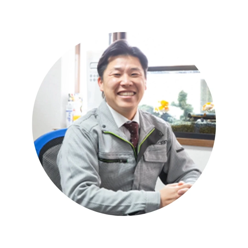 グレーの作業着を着たアジア人男性がオフィスのような場所で明るく笑顔を見せています。背景には金魚鉢が見えます。