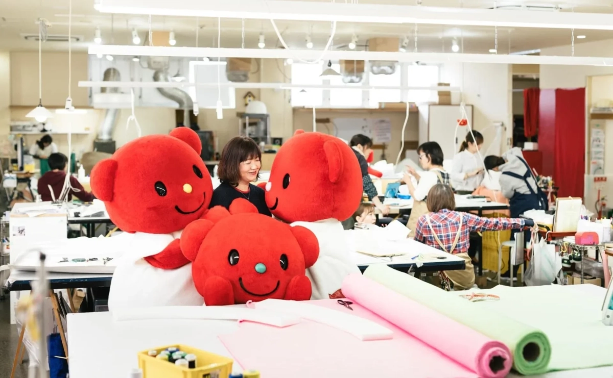ものづくり工房で赤いクマのようなマスコットキャラクターと女性が笑顔で触れ合っている