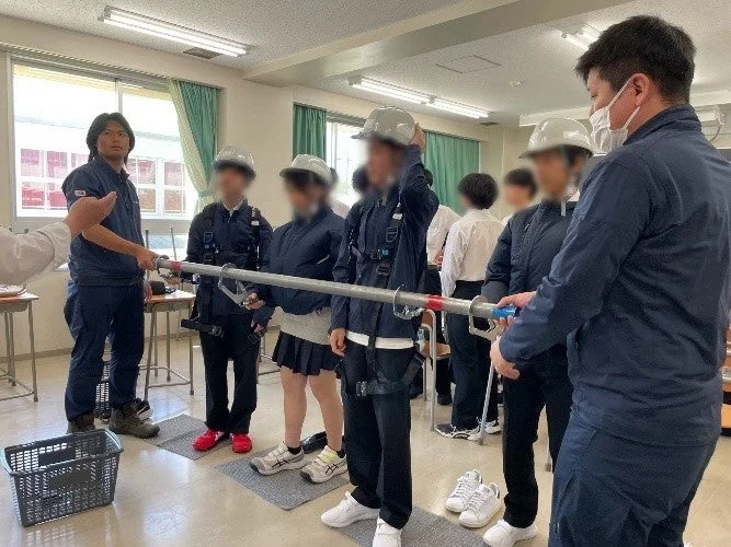 教室で、学生たちがヘルメットと安全帯を着用し、インストラクターから建設現場での安全に関する実習を受けている様子