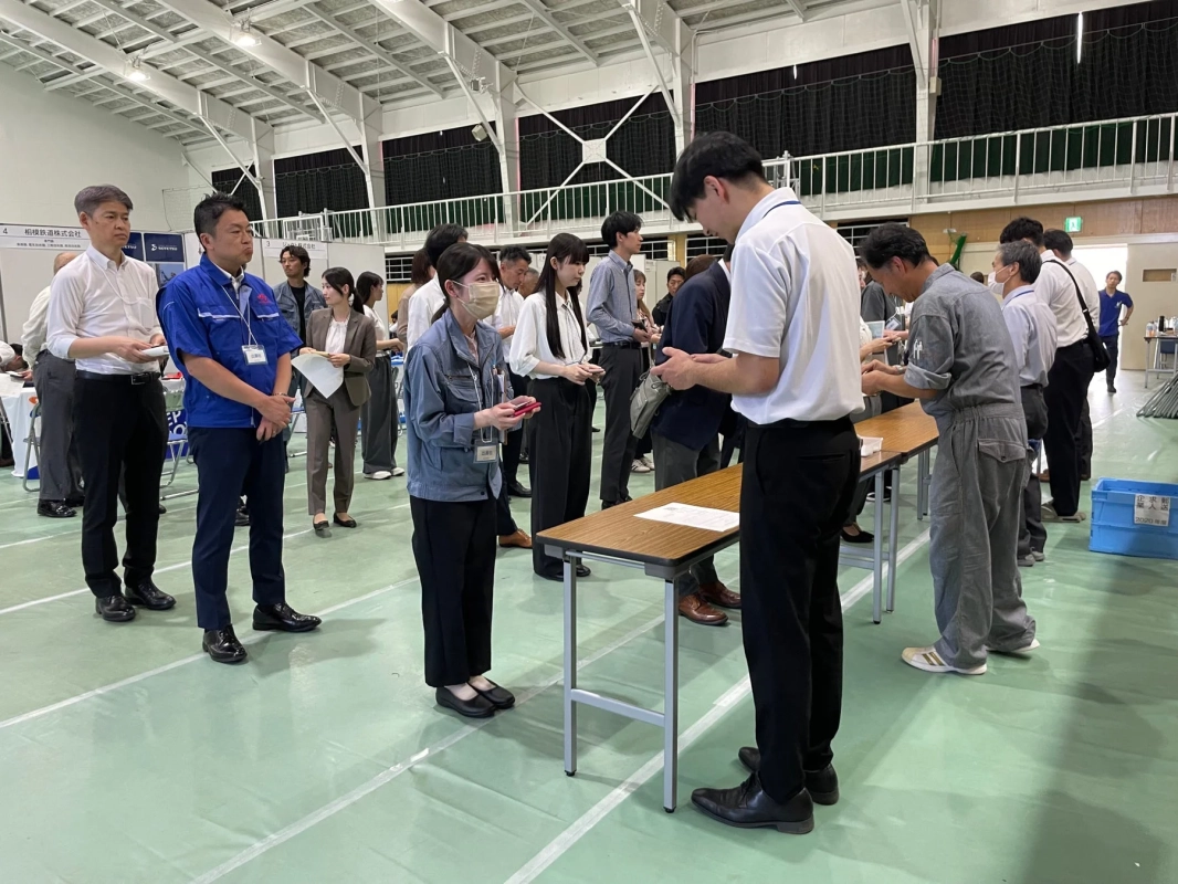 体育館のような広い屋内で、企業のブースや受付のような場所で、参加者が説明を受けたり書類を記入したりしている様子