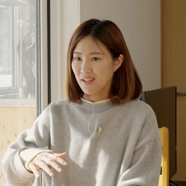 明るい屋内で、ライトグレーのセーターを着たショートヘアの女性が、ラベリアマイクを装着し、手でジェスチャーを交えながら話している様子。窓からの自然光が差し込む中で、和やかな表情を見せています。