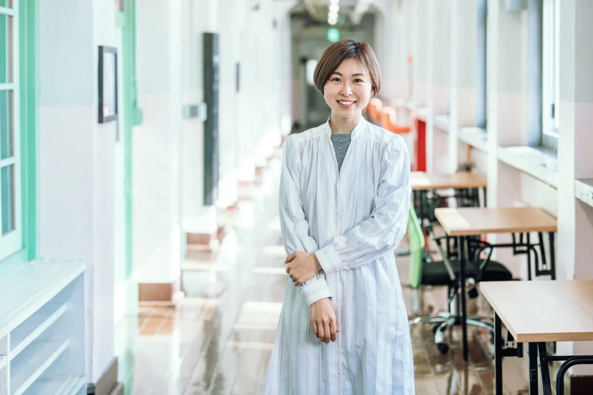 明るい廊下で白いシャツワンピースを着た若い女性が笑顔で立っているポートレートです。学校やオフィスのような清潔感のある空間が背景に広がっています。