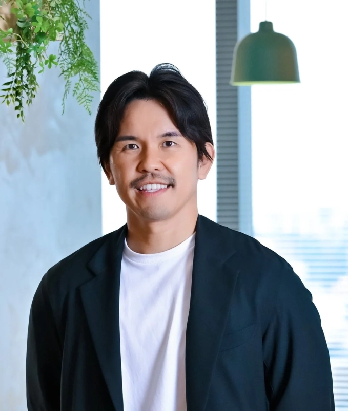 ひげを蓄え、笑顔でカメラを見つめる男性のポートレートです。ダークなブレザーと白いTシャツを着用し、明るい窓と緑の植物、ペンダントライトのある屋内で撮影されています。