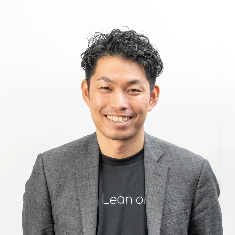 笑顔でカメラを見つめる日本人男性のポートレート。黒いTシャツの上にグレーのジャケットを着用し、くるくるとした髪型が特徴的です。背景は白一色です。