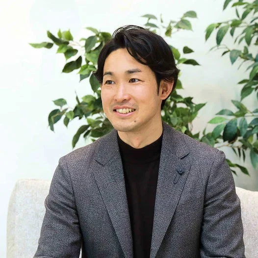 グレーのスーツジャケットと黒のタートルネックを着た日本人男性が、笑顔で左方向を見ています。背景には緑の植物が配置されており、明るく清潔感のある雰囲気です。